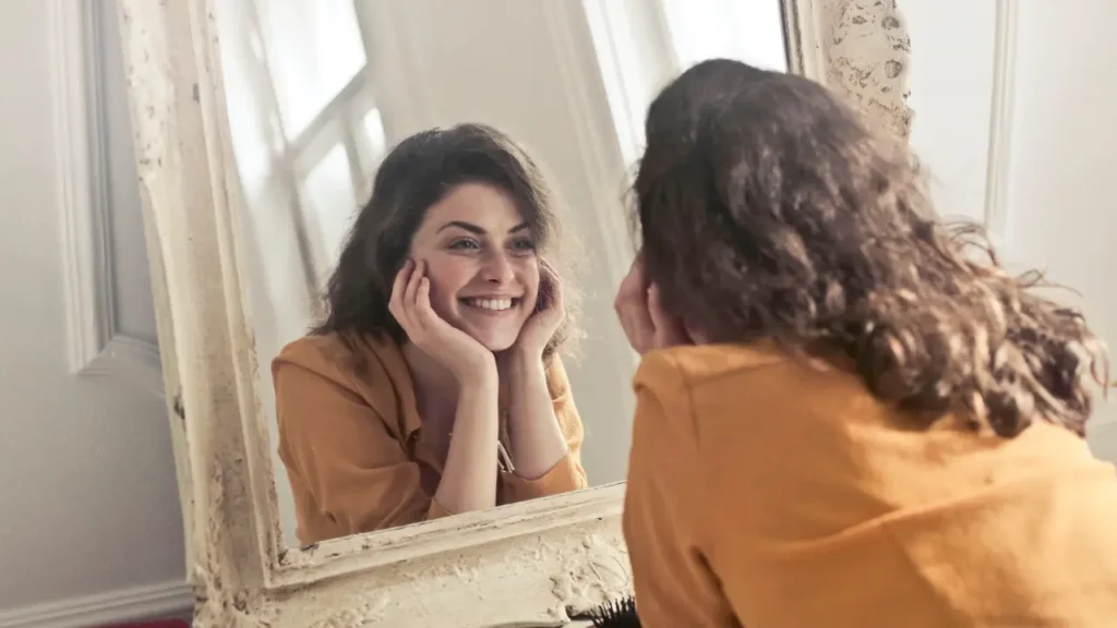 Femme souriante se regardant avec bienveillance dans un miroir, illustrant une estime de soi positive