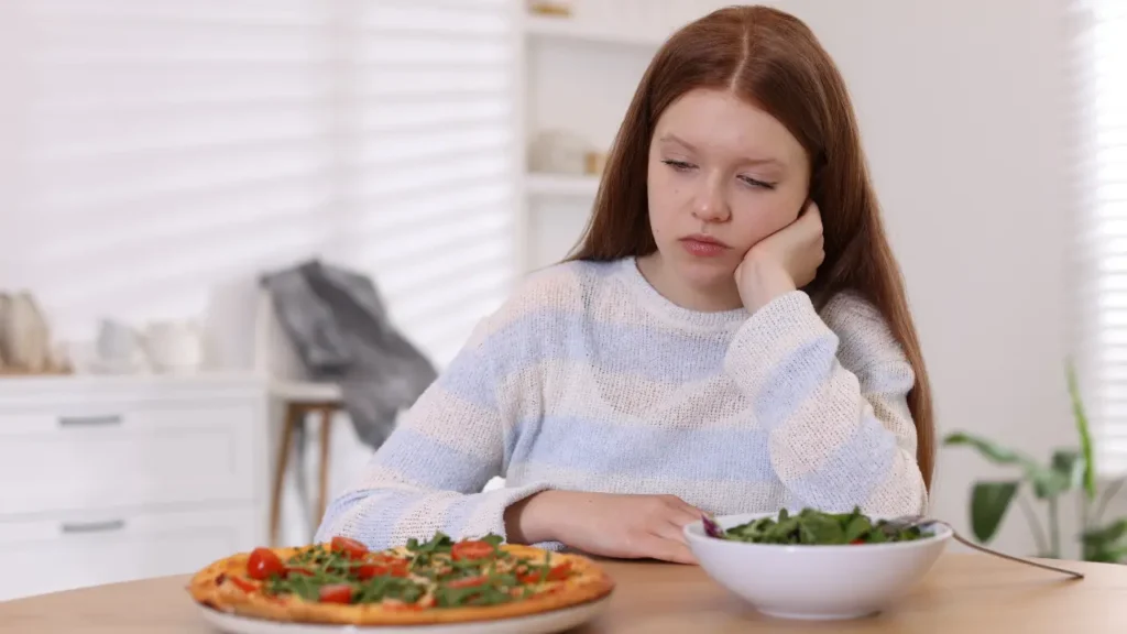 Adolescente pensive devant une assiette de pizza et un bol de salade, illustrant le rapport conflictuel à la nourriture dans les troubles alimentaires